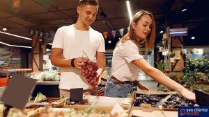A foto mostra duas pessoas escolhendo cachos de uva em um supermercado, tal qual um cliente oculto